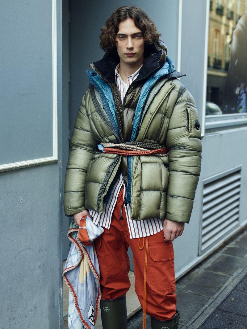 Fashion shot of a male model in olive green puffer jacket with a braided belt, red pants, and olive green boots against urban setting.