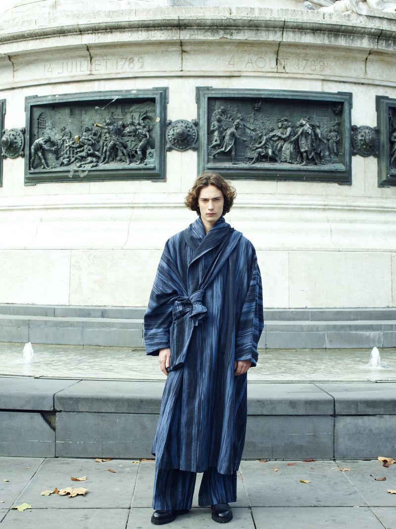 Person wearing blue striped robe and pants standing in front of a monument with bas-relief sculptures and a reflecting pool.