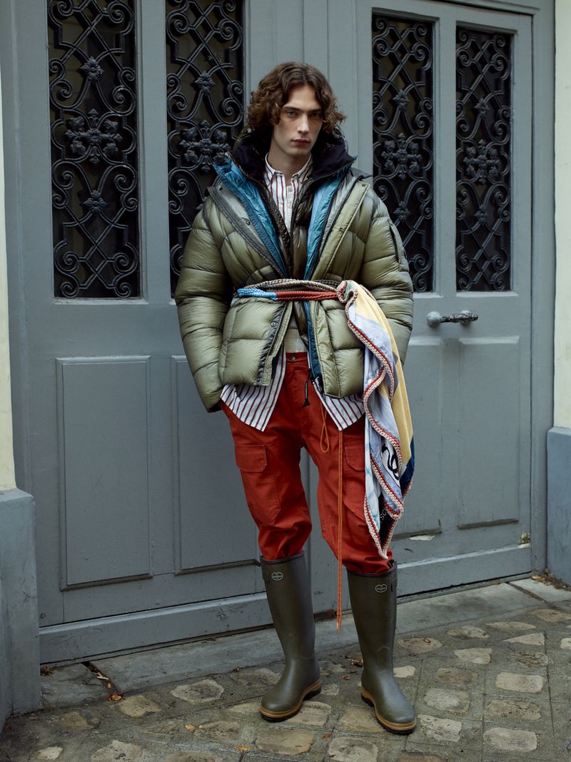 A man poses in a green puffer jacket, striped shirt, and rust-colored pants against a gray door, wearing olive-green wellington boots.