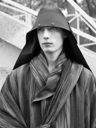 Monochrome portrait of a young adult male wearing a striped kimono-style wrap and a peaked conical hat with buttons on outdoor stairs.