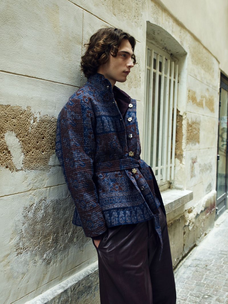 A fair-skinned adult male with curly brown hair poses in an urban alleyway wearing a belted blue patterned jacket and brown trousers. 