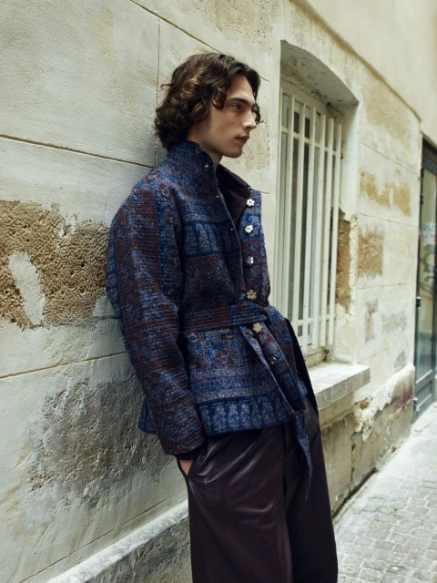 A fair-skinned adult male with curly brown hair poses in an urban alleyway wearing a belted blue patterned jacket and brown trousers. 