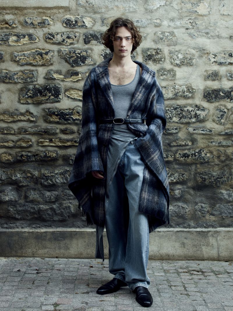 Full-length shot of a male fashion model in a gray top and draped plaid coat standing outdoors against a stone wall backdrop.