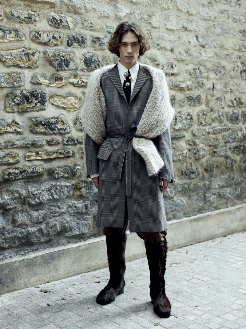 Full shot of androgynous fashion model wearing a grey plaid coat, large off-white knit scarf, and fur boots standing against a stone wall.