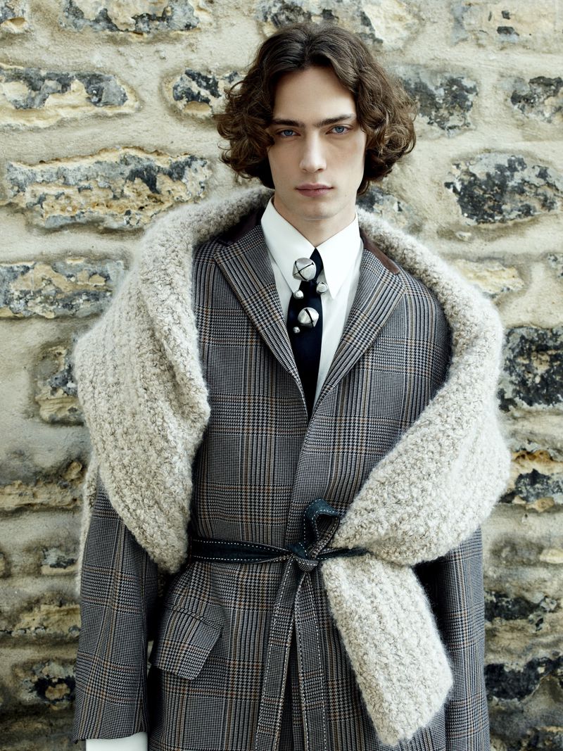 Fashion portrait of a male model wearing a grey plaid suit, white shirt, dark tie, and a textured grey scarf against a stone wall.