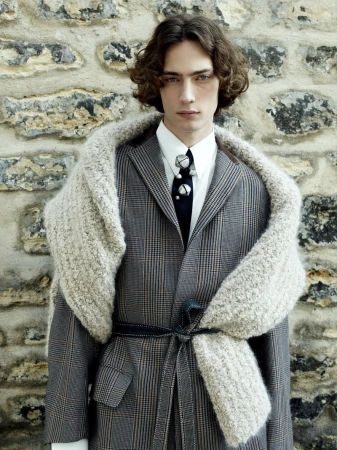 Fashion portrait of a male model wearing a grey plaid suit, white shirt, dark tie, and a textured grey scarf against a stone wall.