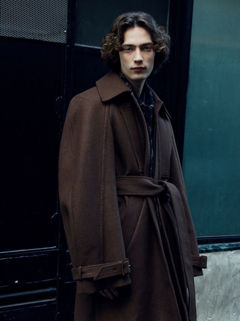 A young adult male stands in a brown overcoat in front of a dark urban background, looking directly at the camera with a serious expression.