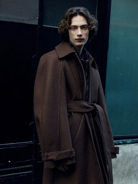A young adult male stands in a brown overcoat in front of a dark urban background, looking directly at the camera with a serious expression.