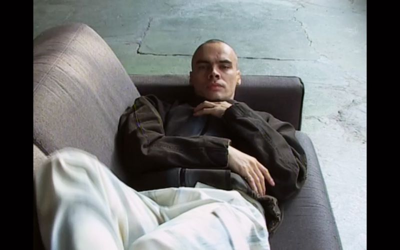 Adult male with a shaved head resting on a gray couch in front of a concrete wall, wearing a brown jacket and light-colored pants.