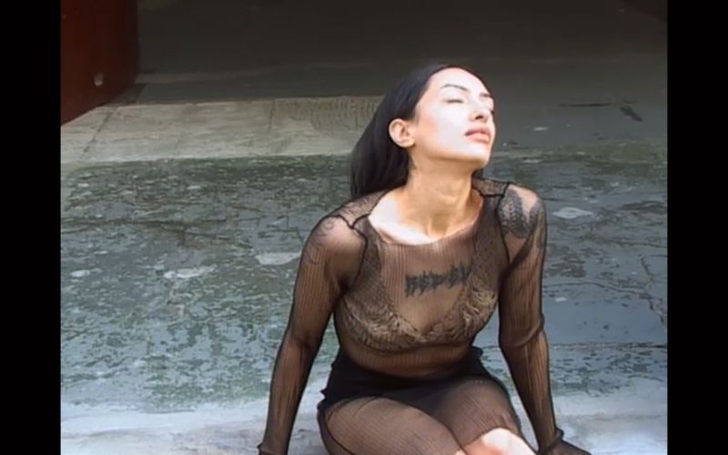 Woman in a sheer long-sleeve mesh top and black skirt sitting on stone pavement with her head tilted back.