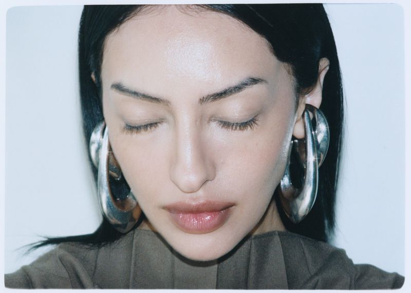 Close-up of a woman with dark hair and large silver earrings, eyes closed, wearing a tan-colored top against a plain white background.
