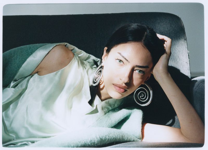 Portrait of a young woman with large spiral earrings, reclining on a couch in soft lighting wearing a light green satin top.