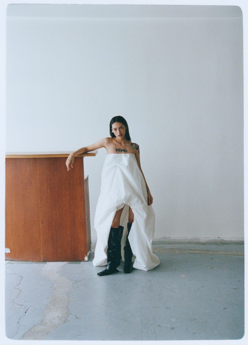 Adult female posing indoors in a white dress and black boots next to wooden cabinet against a plain background.