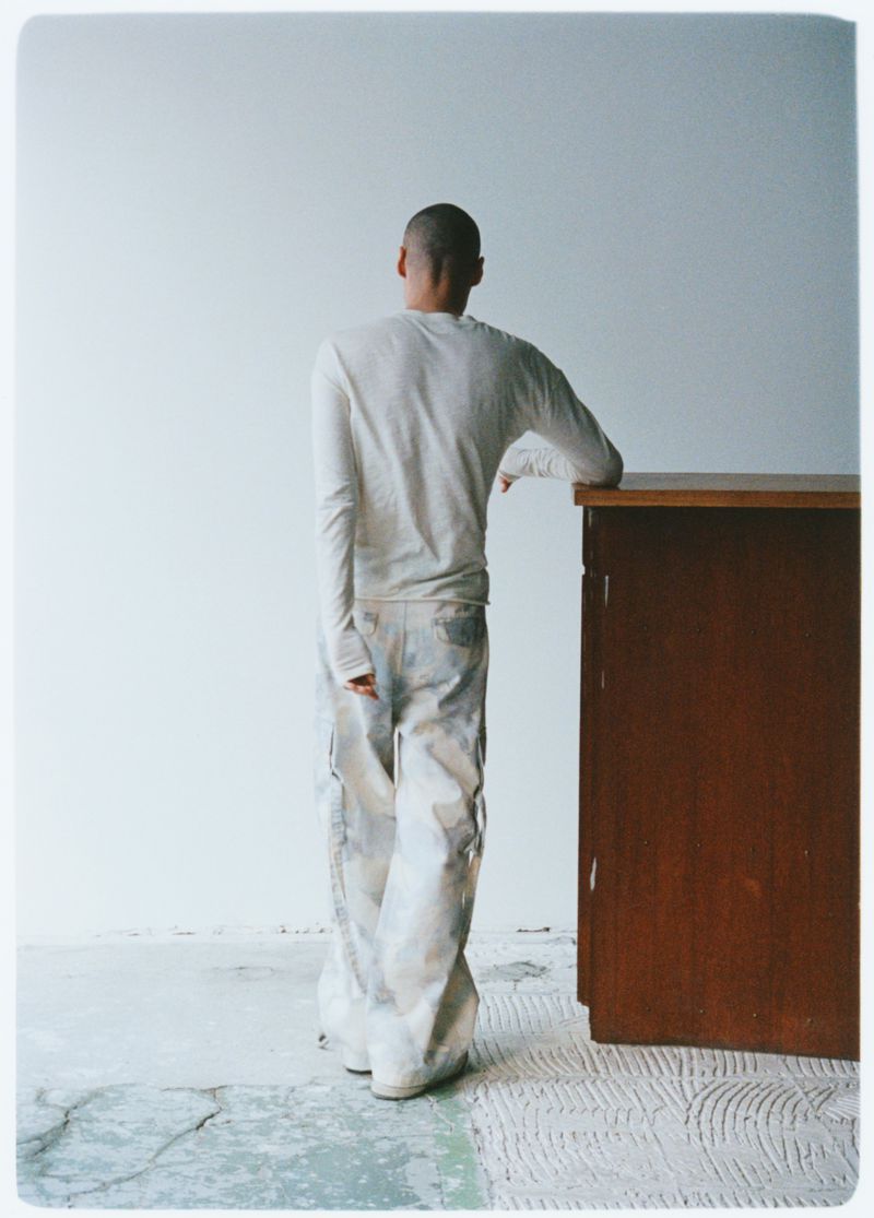 Back view of a person with a shaved head in a long-sleeve shirt and light pants, leaning against a dark brown cabinet in an indoor setting.