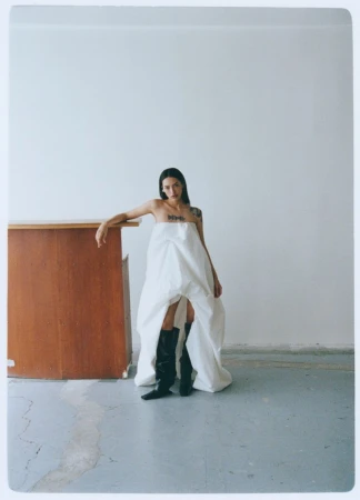 Model in voluminous white dress and black boots leaning on a wooden counter against a white wall.
