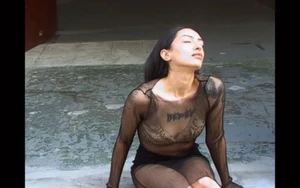 Woman in a sheer long-sleeve mesh top and black skirt sitting on stone pavement with her head tilted back.