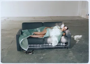 Woman reclining on a dark grey sofa partly draped in a pale green blanket inside a large sparse room.