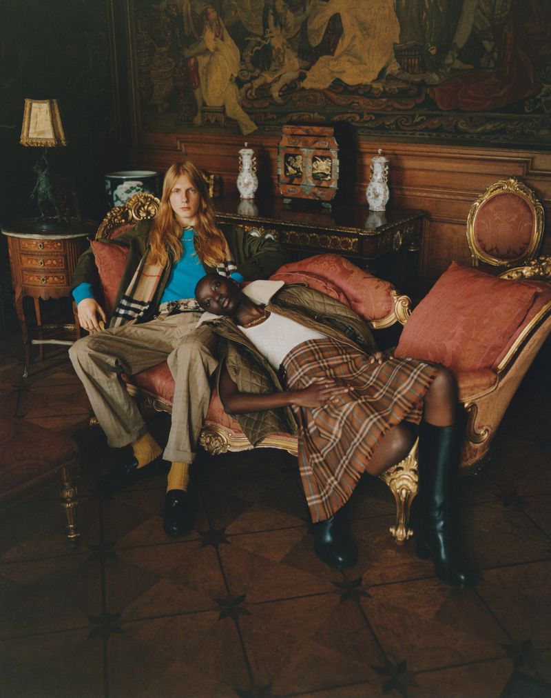 Two people in a room with vintage furniture. One person is sitting and another is reclining on a long red chaise lounge.