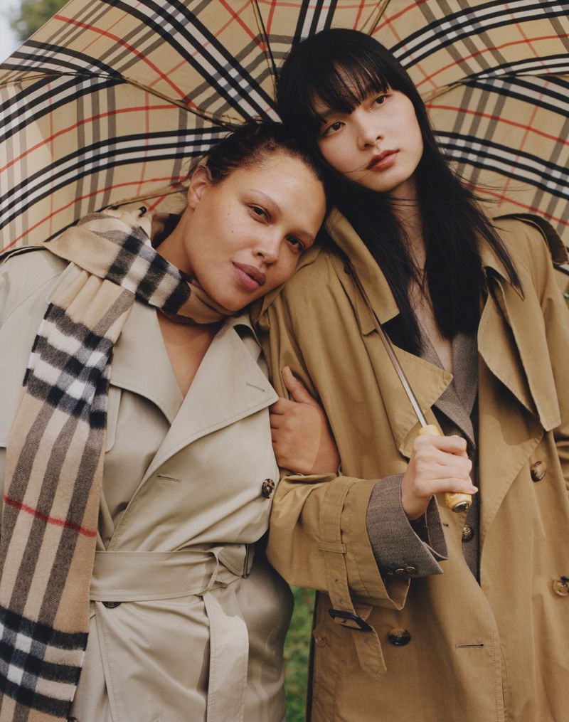 Two diverse women pose closely in trench coats under a Burberry plaid umbrella, set against a muted green background in soft light.