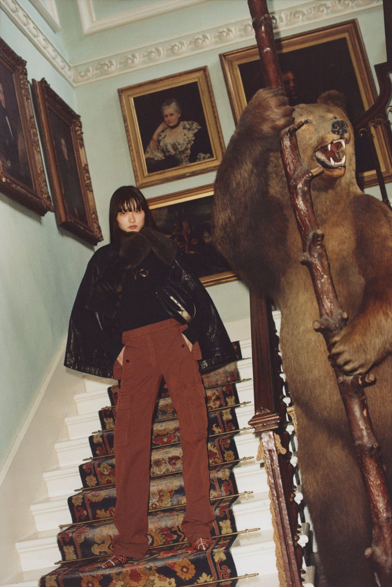 A woman in rust-colored pants and a black top poses on a carpeted staircase with a taxidermied bear and framed artwork in the background.