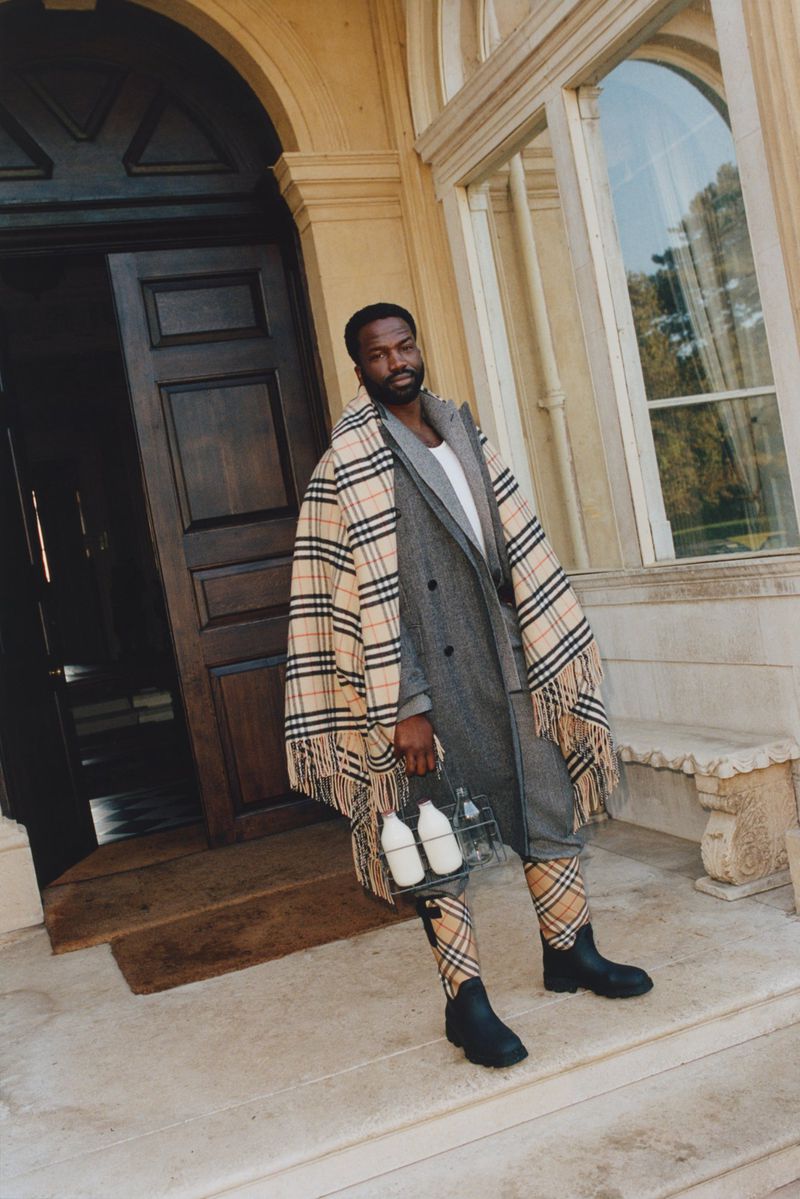 Stylish black male model in plaid coat and matching leggings holds milk bottles outside of stone building on a sunny day.