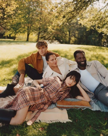 Group of four adults in fashionable clothing, sitting on a blanket in a sunny park surrounded by trees. Lifestyle fashion photography.