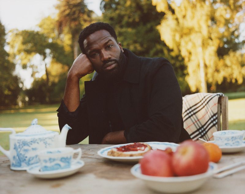 Outdoor portrait of an adult male in a black coat sitting at a table with a tea set, sliced bread with jam, apples and an orange.