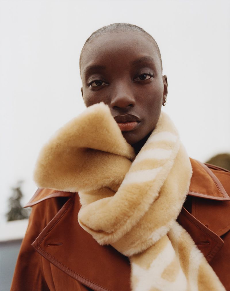 Close-up portrait of a Black woman wearing a brown coat and a striped beige faux fur scarf, against a blurred outdoor background.