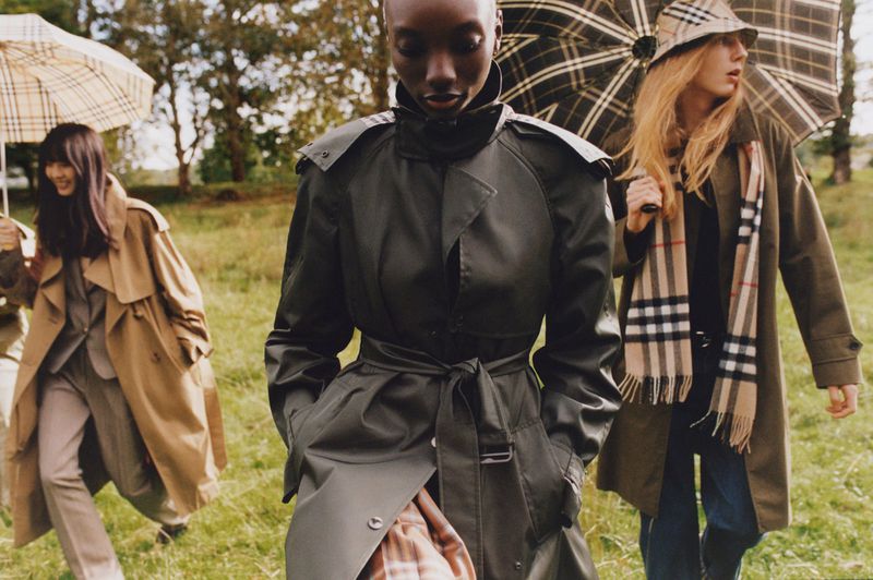 Three models wearing trench coats and plaid accessories walk outdoors holding umbrellas across a grassy meadow.