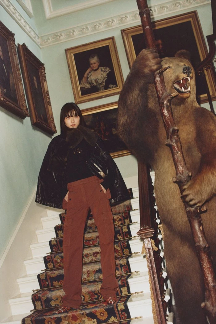 Model in rust-colored trousers and black jacket posing on an ornate carpeted staircase next to a large taxidermy bear.