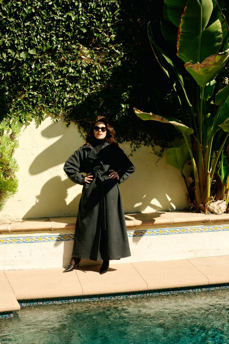Woman in a long belted coat stands next to a pool with a backdrop of greenery, bathed in warm sunlight and showcasing outdoor fashion.