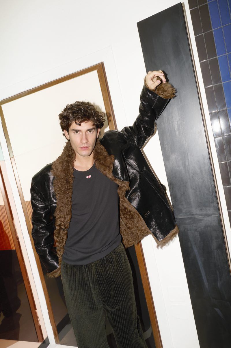 A person wearing a black jacket with fur trim and corduroy pants poses indoors, leaning against a geometric display, in a fashion shot.