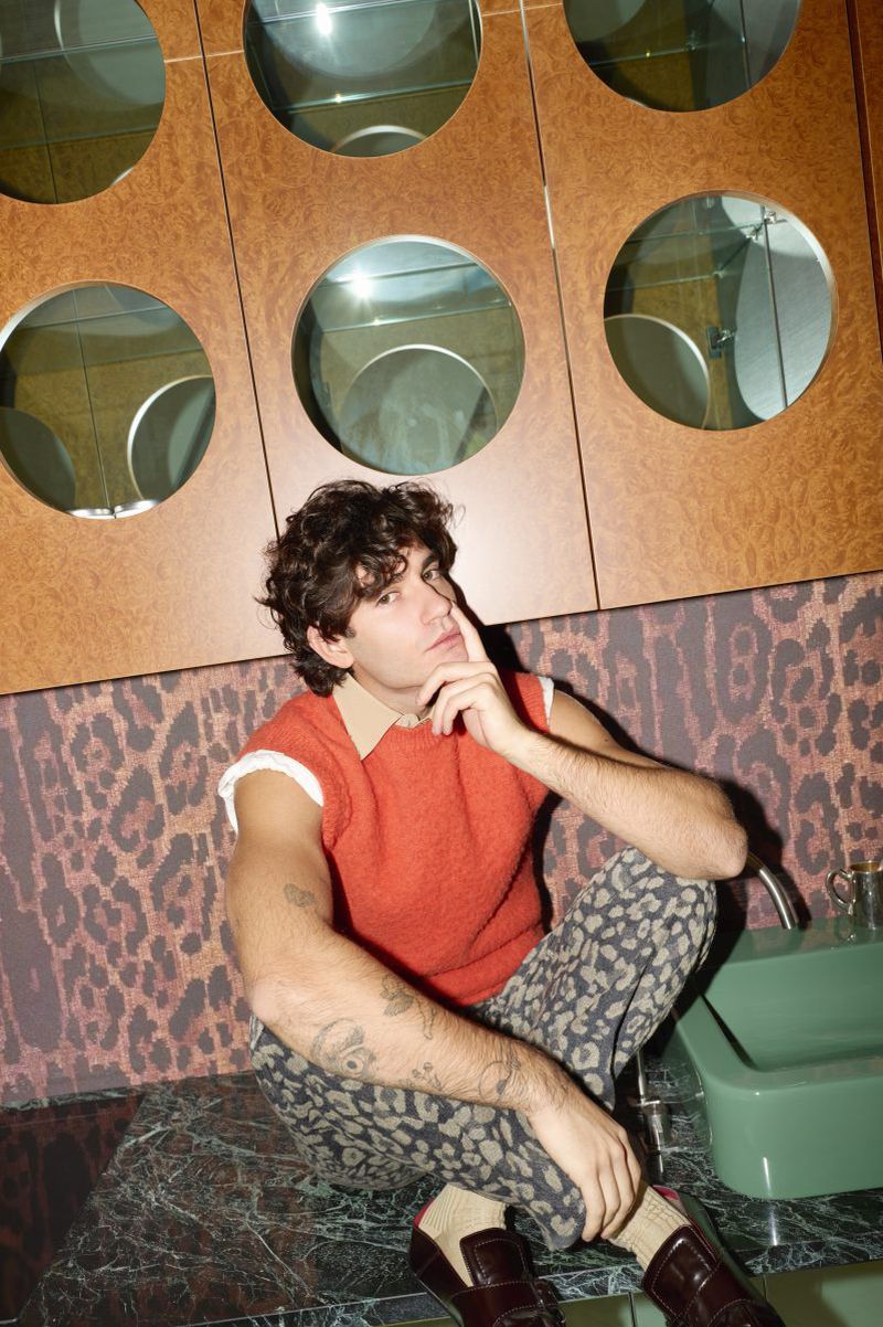 Young man with curly hair sits on a marble countertop wearing an orange top, patterned pants and loafers in an indoor bathroom setting.