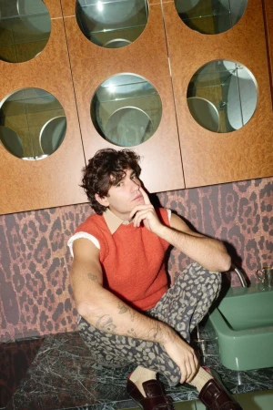 Young man with curly hair sits on a marble countertop wearing an orange top, patterned pants and loafers in an indoor bathroom setting.