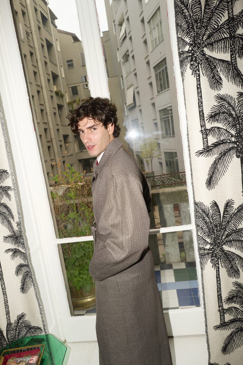 Man in a herringbone coat stands near a window with tropical-patterned curtains, looking toward the viewer. The background shows city buildings.