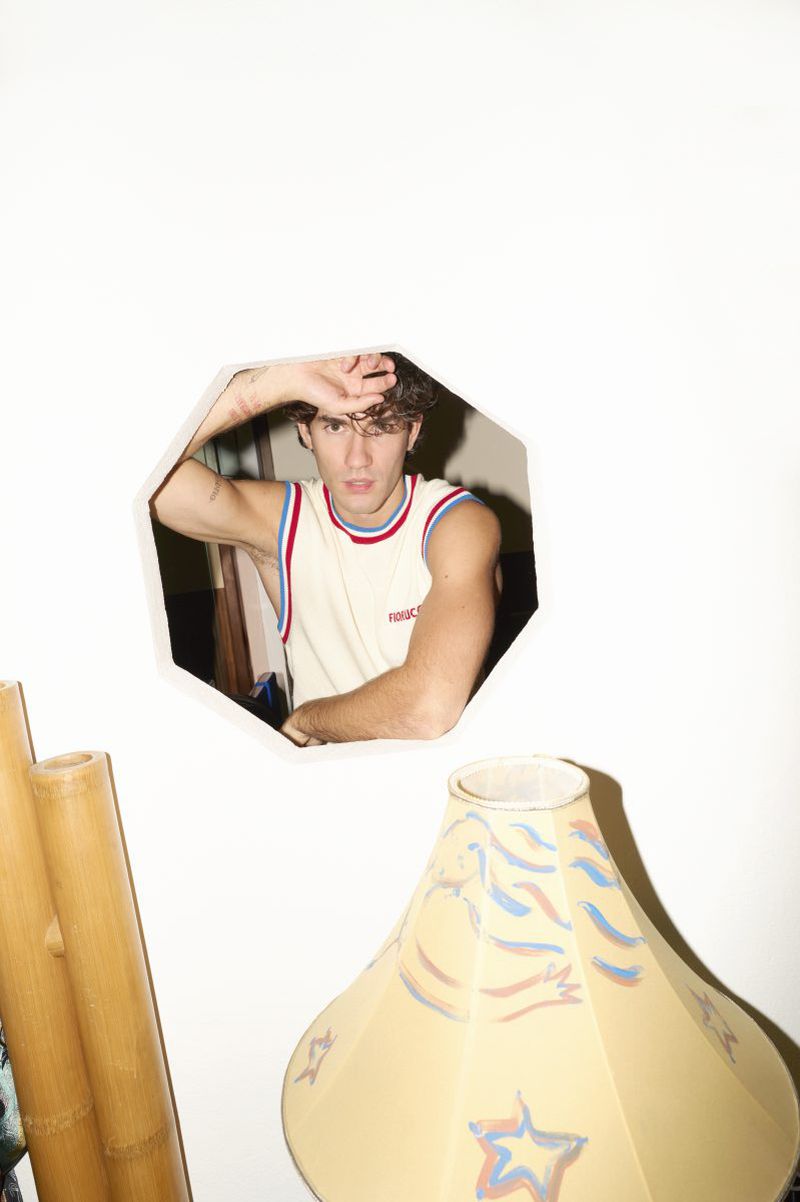 Young man in tank top peeks through octagon wall opening with decorative bamboo and lamp in background indoors. Interior shot.