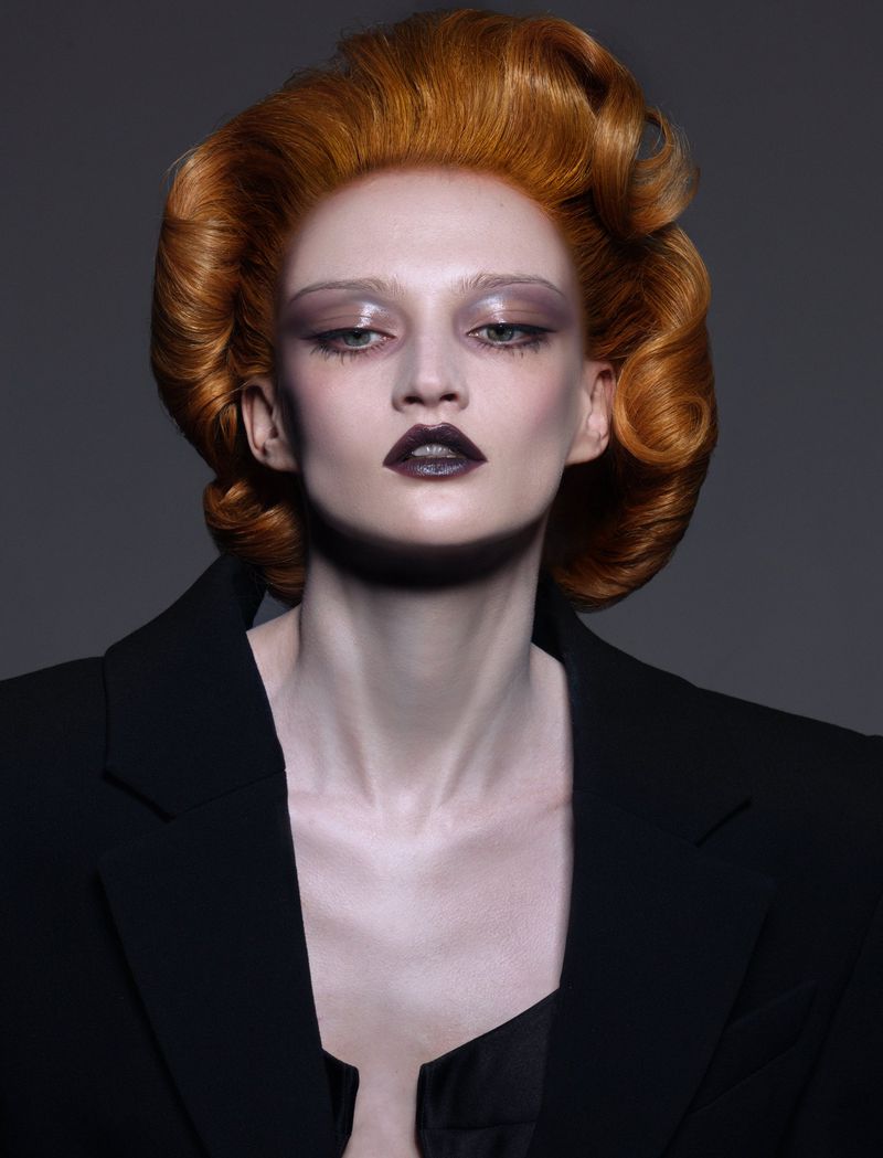 Close-up of a red-haired adult female fashion model wearing a black blazer and dark lipstick against a neutral gray backdrop.