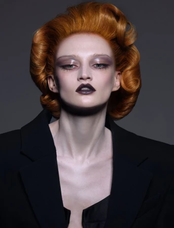 Close-up of a red-haired adult female fashion model wearing a black blazer and dark lipstick against a neutral gray backdrop.