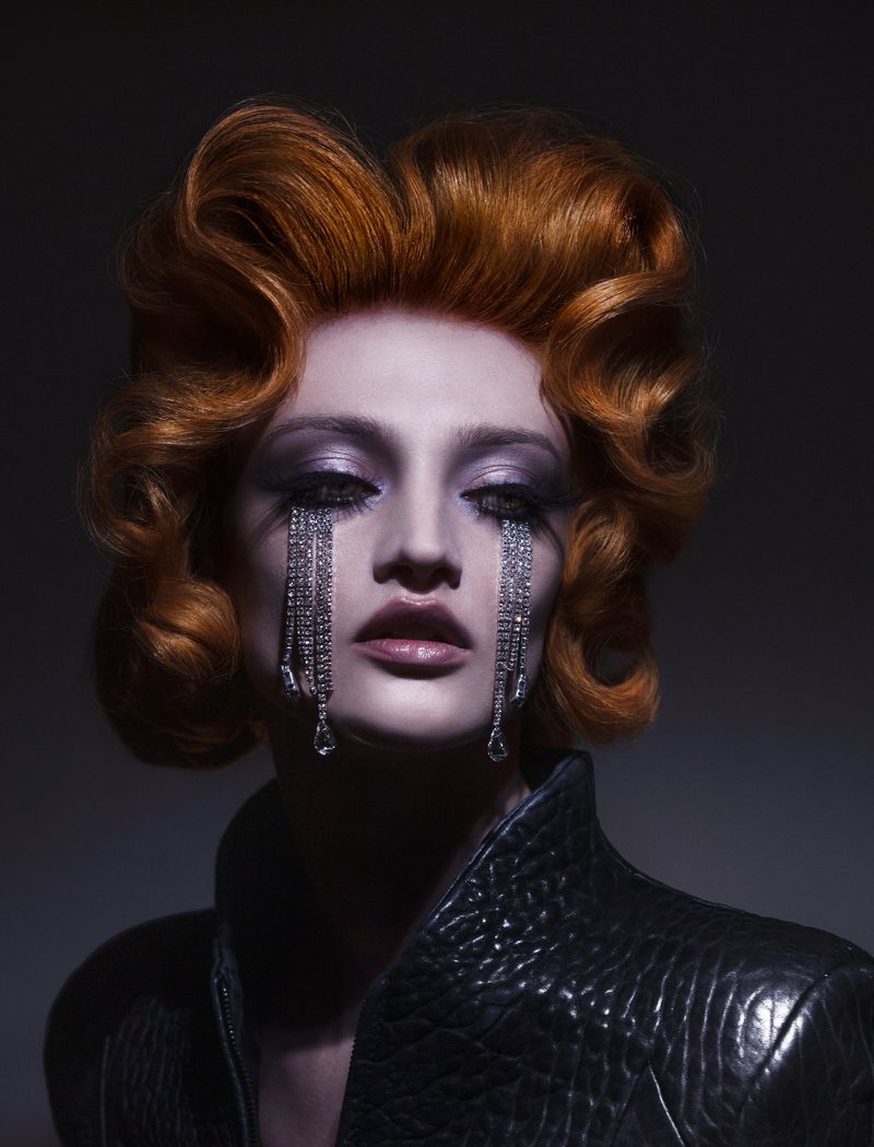 Portrait of a woman with curled red hair, rhinestone eye makeup, and a black jacket, set against a dark studio background.