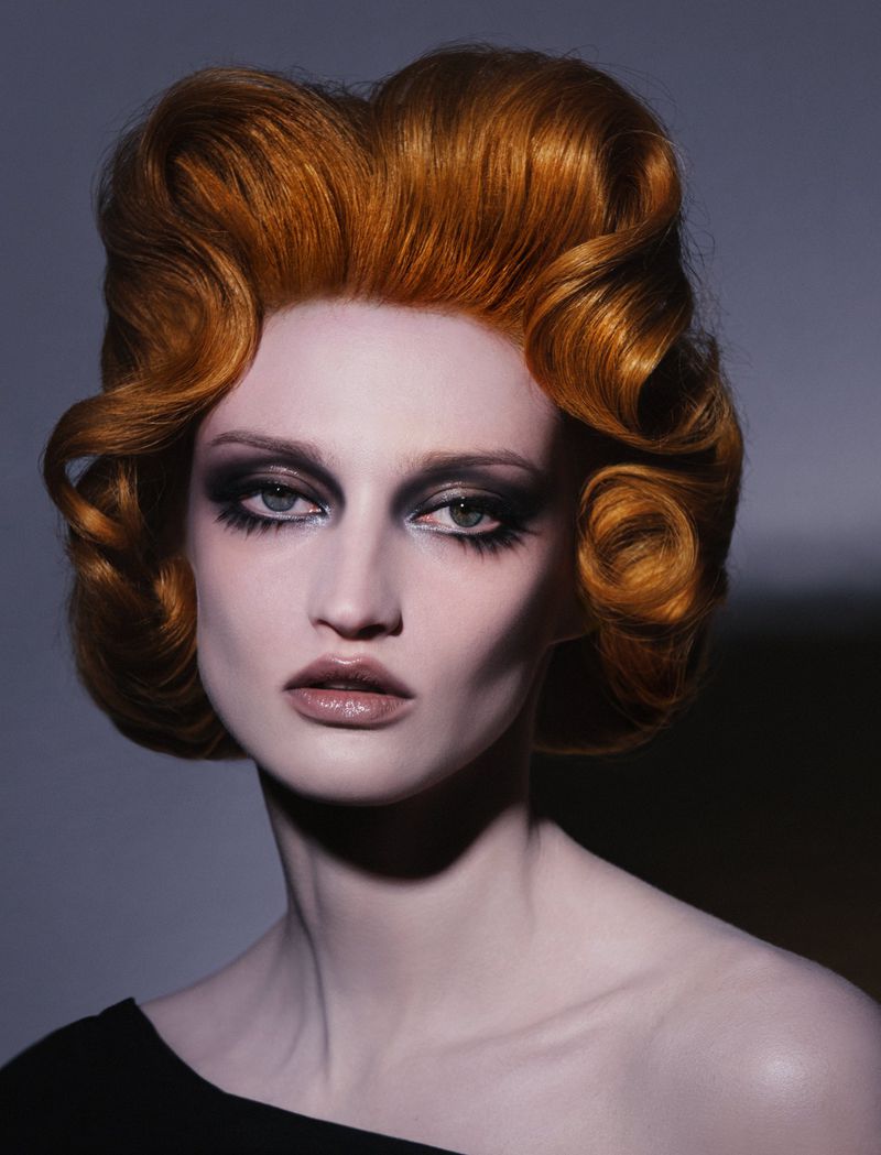 Close-up portrait of an adult woman with red hair styled into voluminous curls, wearing black clothing against a gray background.