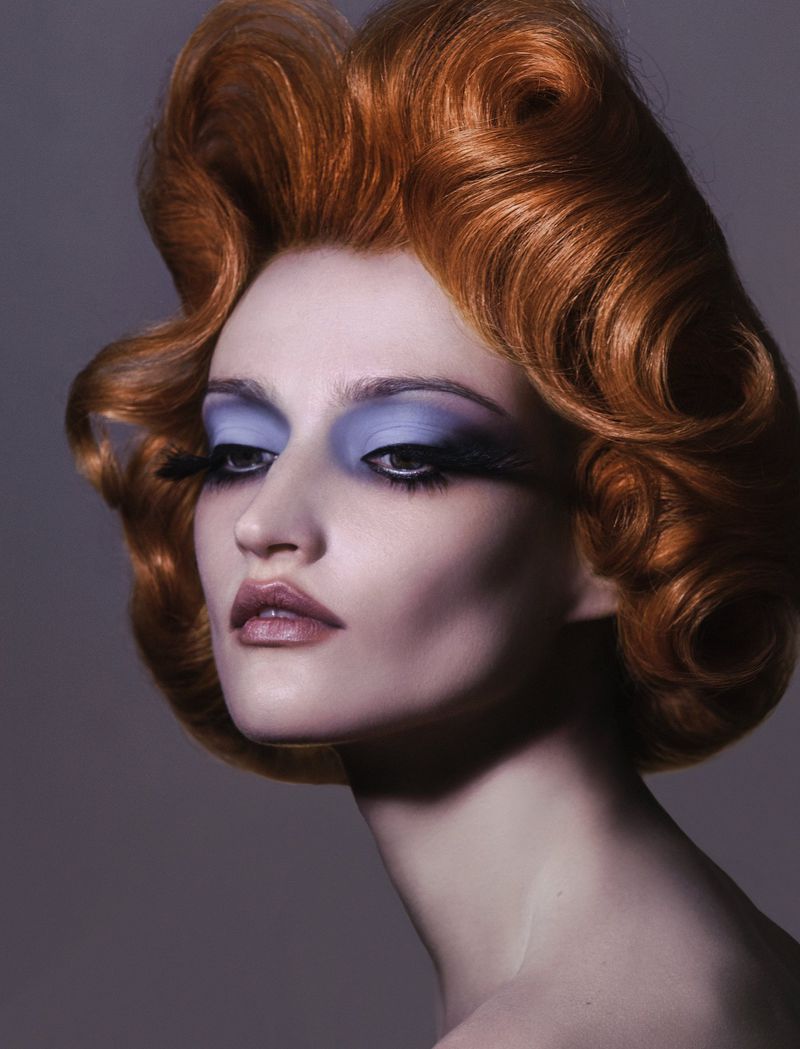 Close-up beauty shot of adult female model with copper-red wavy hairstyle and dramatic lilac eye makeup on a grey gradient background.