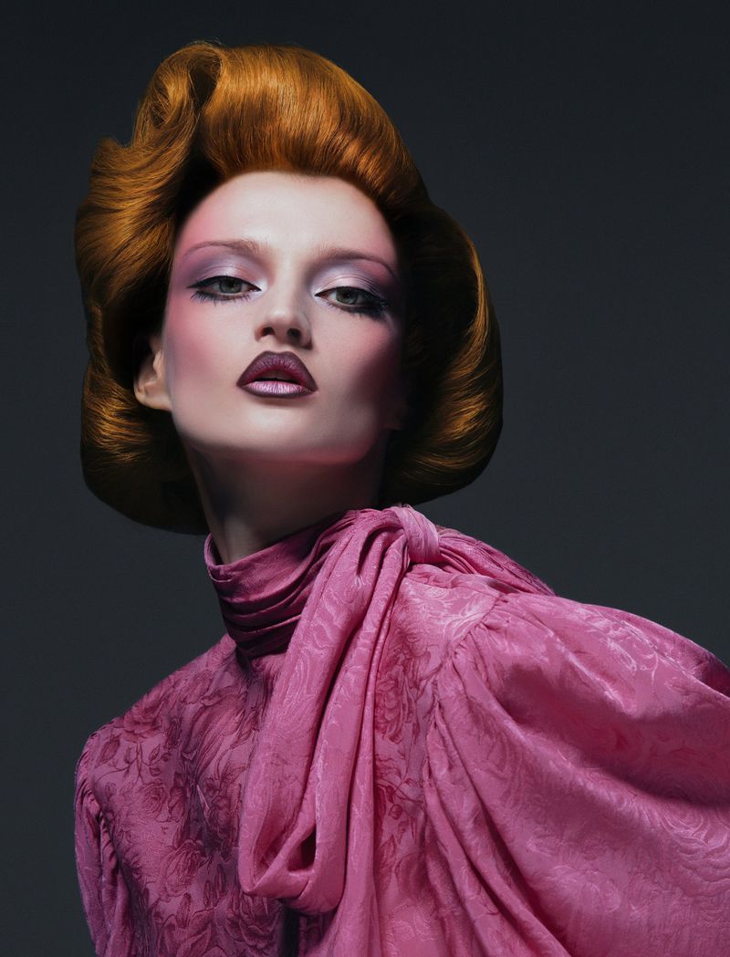 Elegant fashion portrait of a woman with auburn hair and heavy makeup wearing a pink dress in a photo studio setting.