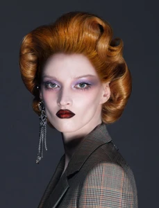 Close-up beauty portrait of a woman with voluminous red hair, purple eyeshadow, dark lips, and a plaid blazer.