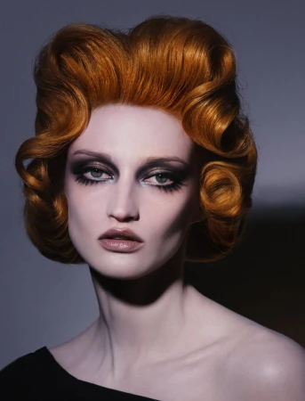 Close-up portrait of a woman with voluminous ginger sculpted hair, heavy dark eye makeup, and pale skin against a grey gradient background