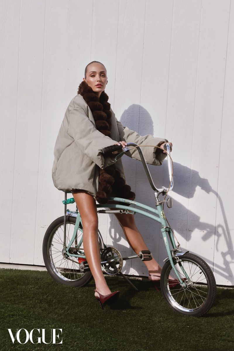 A woman in a coat sits on a blue bicycle outdoors on green grass in front of a white wall, styled in a Vogue fashion editorial.