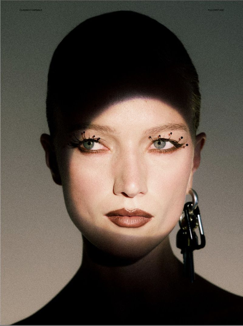 Close-up portrait of a woman with dark lipstick, exaggerated eyelashes, and silver earrings, partially obscured by shadow in a studio setting.