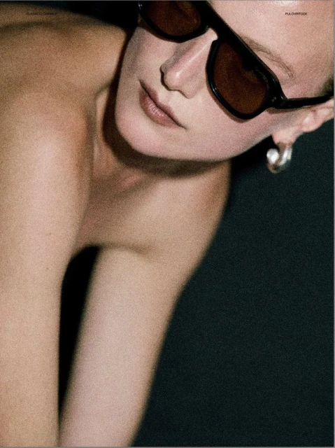 Close-up fashion shot of a fair-skinned adult female model wearing sunglasses and earrings against a dark, blurred background.
