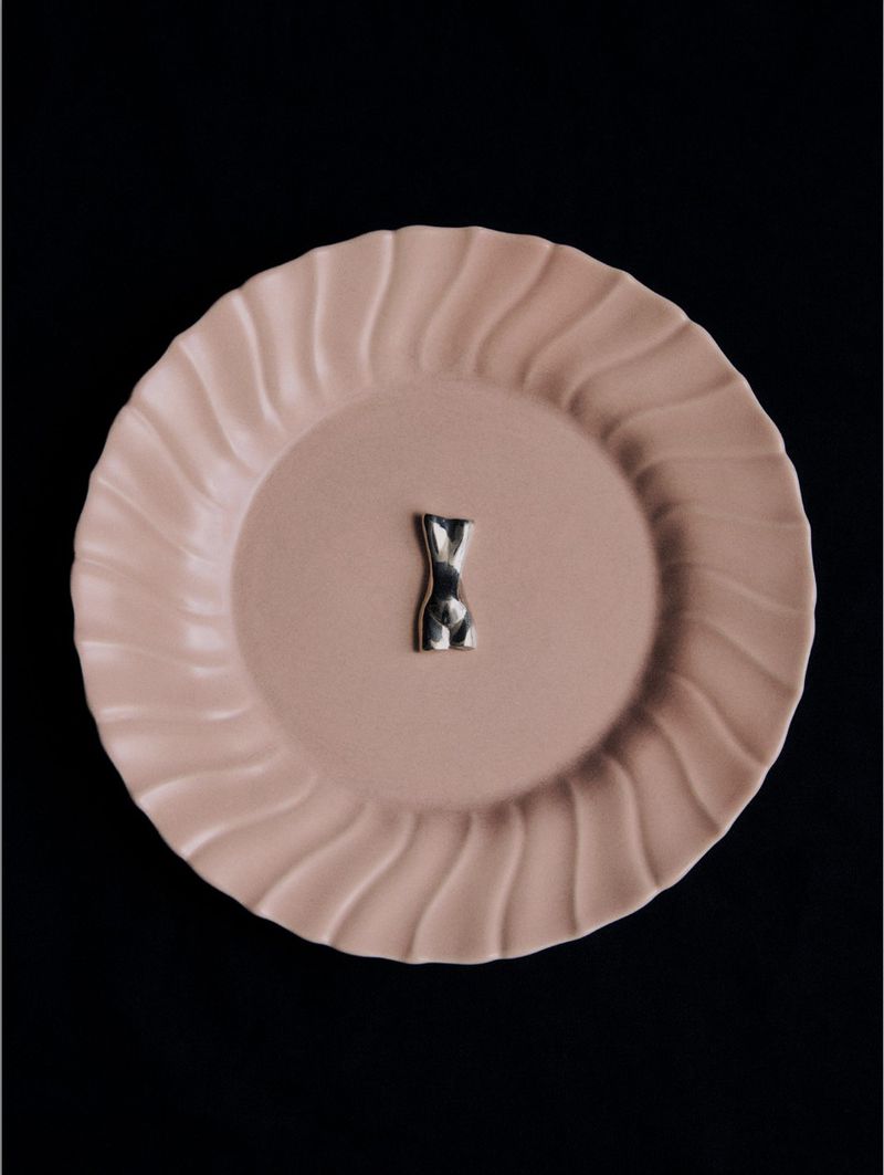 Conceptual still life featuring a small silver torso sculpture centered on a pastel pink plate against a stark black background.