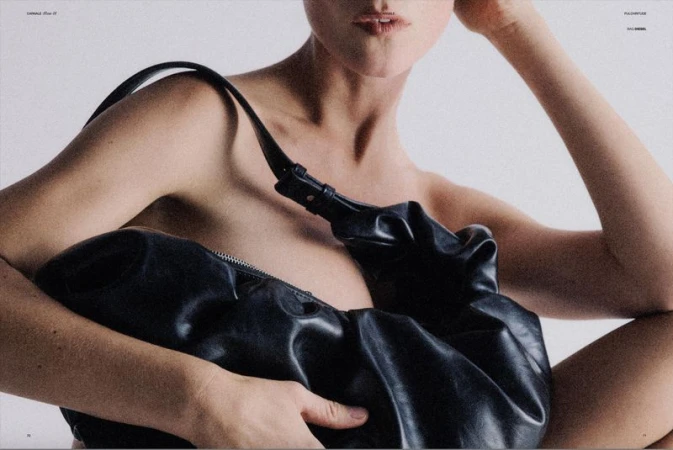 Close-up fashion shot featuring an adult woman with fair skin holding a black leather bag against a plain white background.