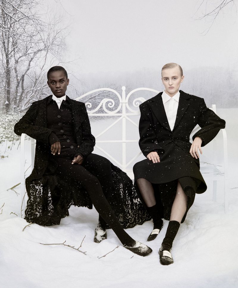 Two fashion models in black outfits pose on a white bench in a snowy outdoor setting with bare trees and soft, diffused lighting.